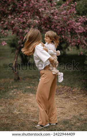 Beautiful mother and daughter against the background of a blooming apple tree. Mom rides her little daughter on her back. Stylish clothes in neutral colors. Mom and daughter having fun
