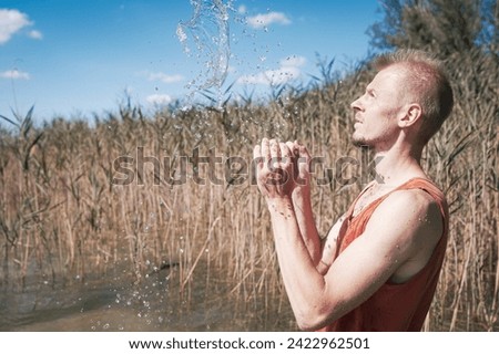A man wearing a muscle shirt stands on the shore of a lake and has scooped water into the air. Royalty-Free Stock Photo #2422962501
