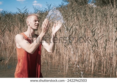 A man wearing a muscle shirt stands on the shore of a lake and has scooped water into the air. Royalty-Free Stock Photo #2422962503