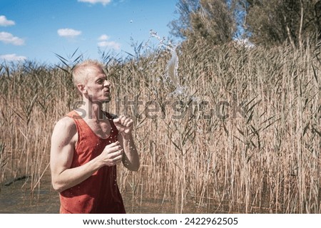 A man wearing a muscle shirt stands on the shore of a lake and has scooped water into the air. Royalty-Free Stock Photo #2422962505