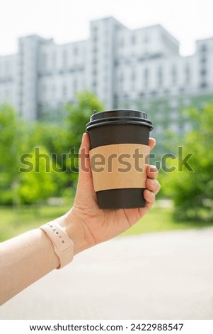 woman's hand holding black hot cup of coffee tea in front of buildings trees and sky
