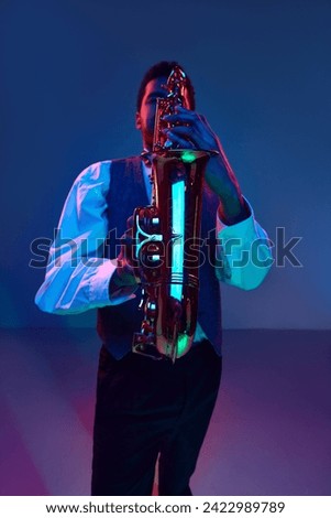 Jazzman. Elegant saxophonist dressed white shirt and vest playing jazz melodies in blue-pink light against gradient studio background. Concept of jazz, blues, classy instrumental music, festivals. Ad