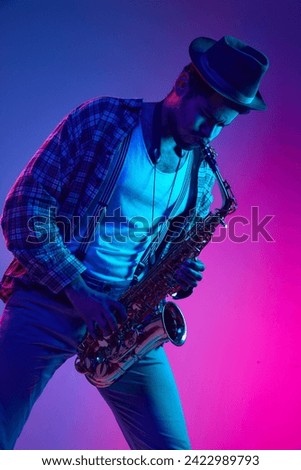 African-American man dressed retro, in hat playing saxophone against gradient pink and blue background in neon light. Concept of jazz, blues, classy instrumental music, festivals and concerts. Ad