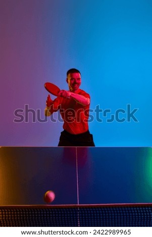 Man in black sport uniform playing table tennis, focused on ball to make perfect serve in neon light against gradient background. Concept of professional sport, healthy lifestyle. Dynamic gel portrait