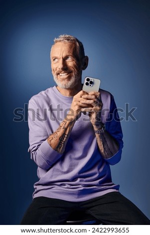 good looking happy mature man with beard in fashionable sweatshirt holding phone and looking away. Translation of tattoo: you have to dig deep wells if you want to get clear water, om, shanti, peace