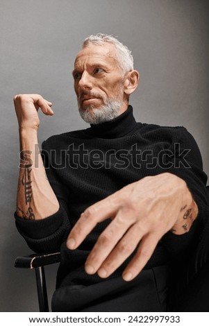 appealing fashionable mature man in stylish turtleneck sitting on folding chair and looking away. Translation of tattoo: om, shanti, peace