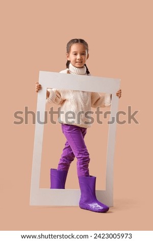 Little Asian girl in rubber boots with frame on beige background