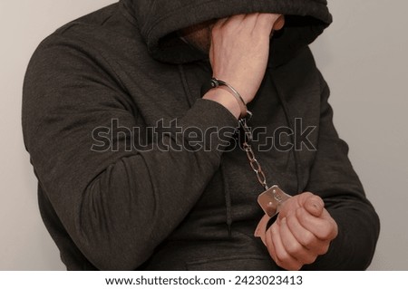 A man with a hood on his head with his hands cuffed hides his face against a gray background.