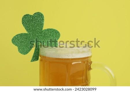 Mug of beer with paper clover on yellow background. St. Patrick's Day celebration