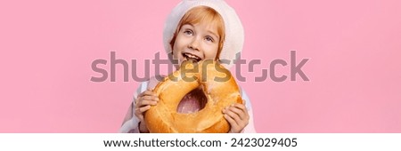 An 8 year old caucasian girl in a bakers hat is holding a large bagel against a pink background. Concepts related to childrens cooking classes or bakery ads. Banner with copy space.
