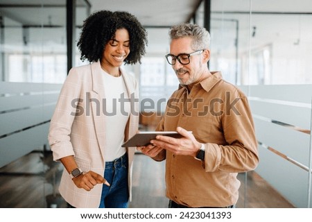 A businesswoman and a senior male colleague collaboratively review content on a tablet in a bright office space, reflecting a multigenerational workplace where experience meets new technology Royalty-Free Stock Photo #2423041393
