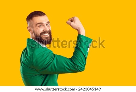 Happy man in suit shows his strong biceps muscles. Cheerful handsome fit bearded young guy in green jacket flexes his arm, raises fist, looks at camera and smiles. Studio portrait, side profile view Royalty-Free Stock Photo #2423045149