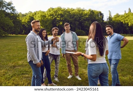Group of happy friends or corporate coworkers standing on grass in summer park on weekend and listening to manager with clipboard listing their tasks and explaining rules of outdoor team building game Royalty-Free Stock Photo #2423045161