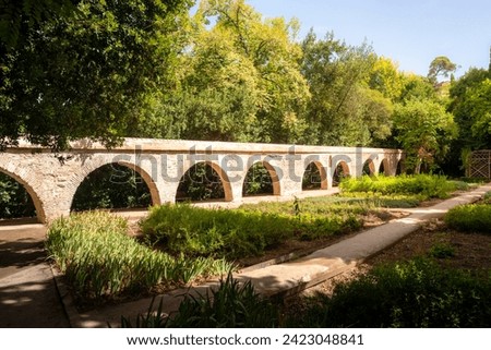 A plant and vegetable garden in warm summer sunlight, covered by trees. Beautiful brick roman aqueduct water moat or wall surrounds pathways and raised beds. blue sky. Vegetation, agriculture.