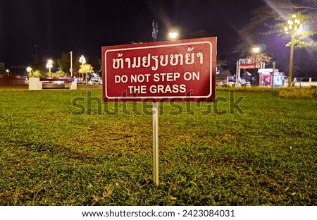 A bilingual "Do Not Step on the Grass" sign prominently displayed in a public park at night, with illuminated street lights in the background
