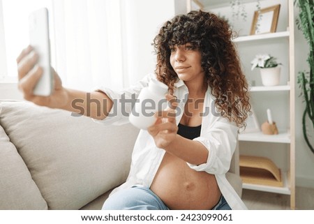 Pregnant woman blogger with a jar of pills and vitamins sits on the couch at home and takes pictures of herself on the phone, maintaining pregnancy and replenishing vitamins and minerals