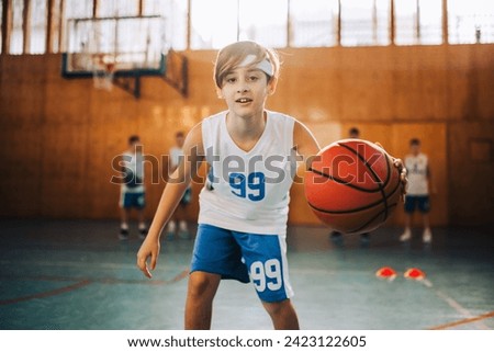 Dynamic portrait of a junior athlete dribbling a ball towards camera and practicing at indoor court. There are his teammates in a blurry background. A young basketball player playing basket on court. Royalty-Free Stock Photo #2423122605