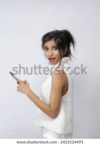 Surprised young Asia lady using mobile phone with positive expression, smile broadly, dressed in casual clothing and looking at camera on white background. Happy adorable glad woman rejoices success