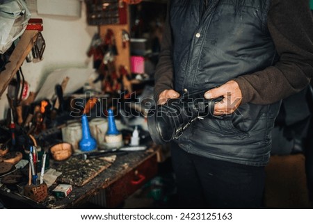 An unrecognizable artisan holding boots for repair while standing at shoemaker workshop. Cropped picture of an old shoemaker holding boots in his shop and preparing for repairing. Small business.