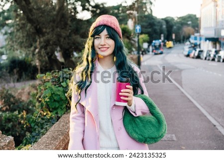 Stylish young smiling hipster woman with color hair walking on street in pink outfit with reusable coffee cup wearing coat, knitted hat, fur bag, happy mood, seasonal fashion, Barbiecore style trend Royalty-Free Stock Photo #2423135325