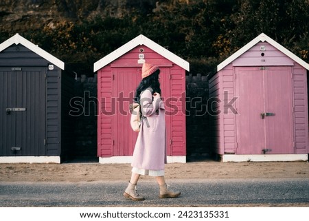 Stylish hipster woman with color hair in pink outfit and backpack walking along wooden beach huts on seaside. Off season Travel concept. Seasonal street fashion. Barbiecore style. Simple pleasures Royalty-Free Stock Photo #2423135331