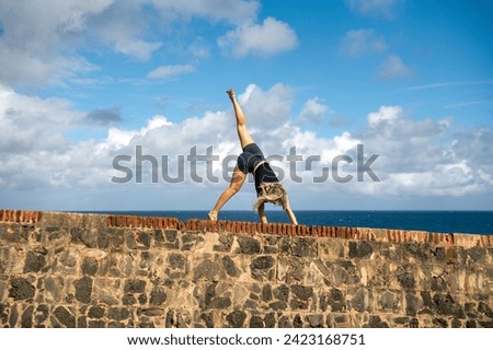 A young blonde woman does a cartwheel and handstand on a stone wall along the sea.