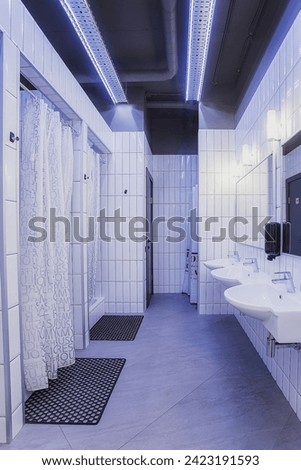 common shower room with shower stalls, white sinks, two mirrors, white tiles. With two black rugs, white curtains on the shower stalls, dark black ceiling and dark tiles on the floor