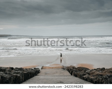 Ramp to a vast sandy beach and rough ocean water, silhouette of a person walking along shows the scale of the place. Lahinch, county Clare, Ireland. Dark dramatic sky. Rough nature scene.