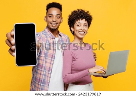 Young smiling couple two friends family IT man woman wear purple casual clothes together hold work on blank screen laptop pc computer use mobile cell phone isolated on plain yellow orange background