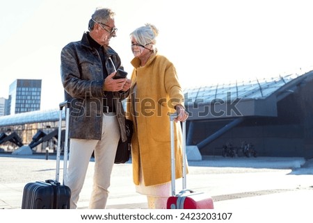 Seniors couple using mobile phone on the train station. Two mature people wearing protective masks. Tourist on the city break. Travel and relationship concept during a pandemic. Outdoor photo.