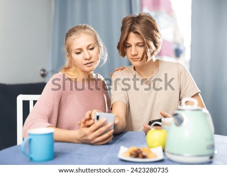 Positive teenager showing pictures on smartphone to his interested mother while sitting at table in cozy dining room