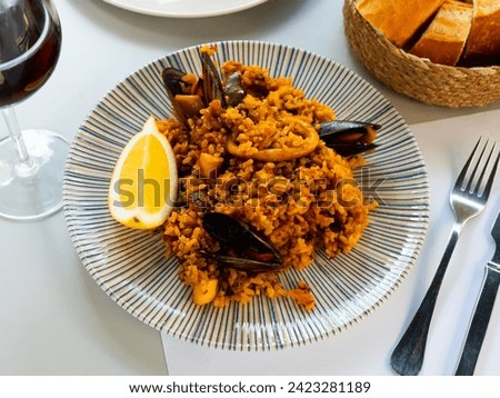 Appetizing racy seafood paella with mussels and squid rings served with lemon slice and glass of red wine. Traditional Spanish cuisine. Royalty-Free Stock Photo #2423281189