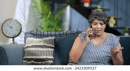Upset Indian old grey-haired woman sitting on sofa couch talking on mobile phone answer unpleasant call received bad shocking news. Disappointed worried senior grandmother feel sad alone indoor home Royalty-Free Stock Photo #2423309527