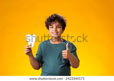 Portrait of child boy holding papercraft tooth and showing thumb up gesture, over yellow background. Dental health concept