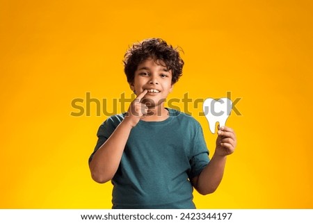 Portrait of child boy holding papercraft tooth and pointing finger at mouth, over yellow background. Dental health concept