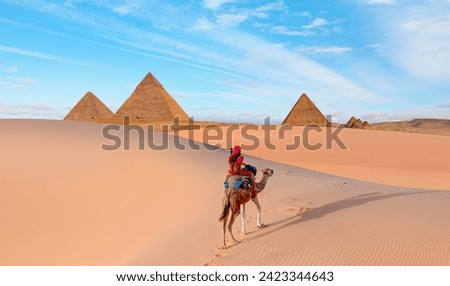 Camels in Giza Pyramid Complex - A woman in a red turban riding a camel across the thin sand dunes - Cairo, Egypt Royalty-Free Stock Photo #2423344643