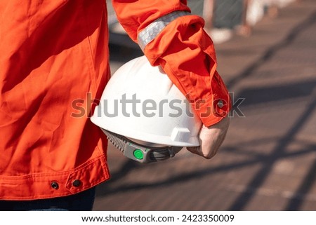An engineer or construction worker is holding a white safety helmet, wearing orange coverall, standing on the working platform walkway. Ready to work in the challenge workplace concept scene.  Royalty-Free Stock Photo #2423350009