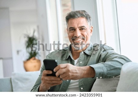 Smiling happy mature middle aged man holding cell mobile phone using smartphone sitting at home on couch, scrolling social media, checking financial apps, buying online, looking at camera. Portrait
