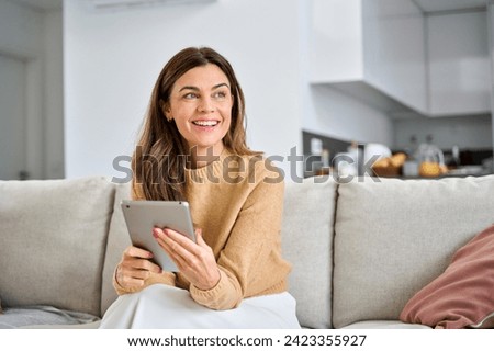 Happy mature older woman using digital tablet sitting on couch at home. Smiling middle aged 45 years old woman looking away at copy space holding tab computer relaxing on sofa in living room. Royalty-Free Stock Photo #2423355927