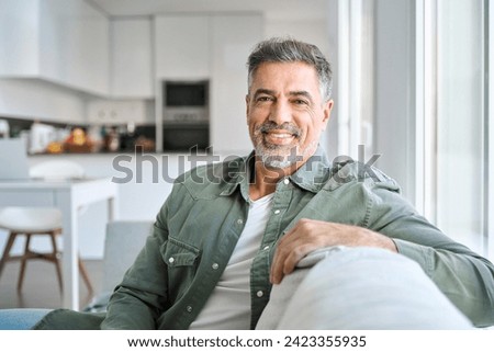 Portrait of happy smiling middle aged mature senior 50 years old bearded man wearing green shirt sitting on couch at home interior looking at camera relaxing on sofa in modern house. Royalty-Free Stock Photo #2423355935