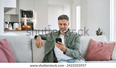 Relaxed happy older mature middle aged man holding mobile phone using cellphone sitting at home on sofa in modern living room, chatting online, texting messages, reading news, shopping in apps. Royalty-Free Stock Photo #2423355957