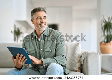 Happy middle aged man using digital tablet relaxing on couch at home. Mature male user holding tab computer holding pad technology device sitting on sofa in living room looking away. Copy space. Royalty-Free Stock Photo #2423355971