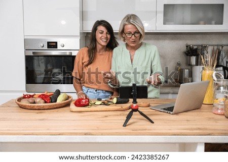 Senior woman cancer survivor and her daughter live streaming vlog via laptop webcam in the kitchen cooking healthy vegetable vegan food for better health and body regeneration after sickness.