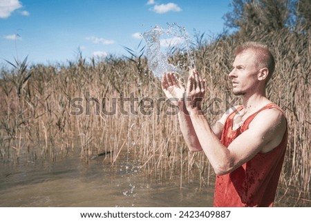 A man wearing a muscle shirt stands on the shore of a lake and has scooped water into the air. Royalty-Free Stock Photo #2423409887