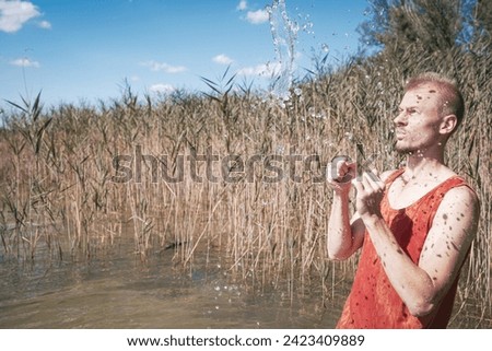 A man wearing a muscle shirt stands on the shore of a lake and has scooped water into the air. Royalty-Free Stock Photo #2423409889