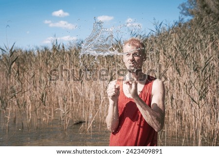 A man wearing a muscle shirt stands on the shore of a lake and has scooped water into the air. Royalty-Free Stock Photo #2423409891