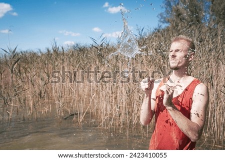 A man wearing a muscle shirt stands on the shore of a lake and has scooped water into the air. Royalty-Free Stock Photo #2423410055