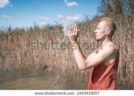 A man wearing a muscle shirt stands on the shore of a lake and has scooped water into the air. Royalty-Free Stock Photo #2423410057