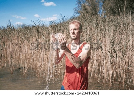 A man wearing a muscle shirt stands on the shore of a lake and has scooped water into the air. Royalty-Free Stock Photo #2423410061