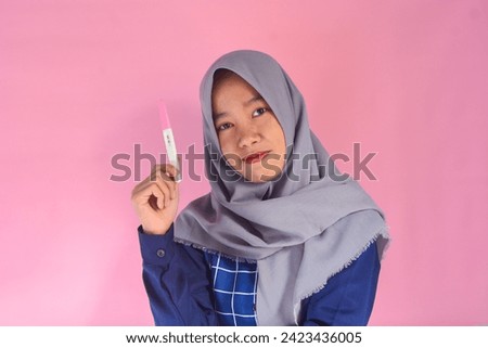 Happy and grateful young woman wearing blue shirt with gray hijab showing her pregnancy test, hand on chest smiling isolated on pink background